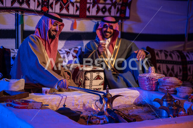 تحريك الجمر في الموقد الطيني باستخدام الملقط، كشتة بر شبابية داخل خيمة تراثية، عادات وتقاليد سعودية في تقديم واعداد القهوة العربية وكرم الضيافه، ارتداء الازياء الوطنية، صديقان عربيان خليجيان سعوديان يرتديان الشماغ والبشت يتبادلان الاحاديث في جلسات شعبية