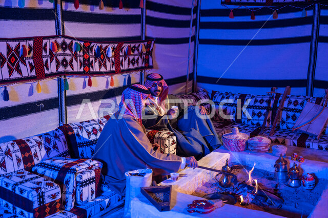 تحريك الجمر في الموقد الطيني باستخدام الملقط، كشتة بر شبابية داخل خيمة تراثية، عادات وتقاليد سعودية في تقديم واعداد القهوة العربية وكرم الضيافه، ارتداء الازياء الوطنية، صديقان عربيان خليجيان سعوديان يرتديان الشماغ والبشت يتبادلان الاحاديث في جلسات شعبية