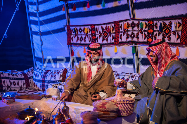 تحريك الجمر في الموقد الطيني باستخدام الملقط، كشتة بر شبابية داخل خيمة تراثية، عادات وتقاليد سعودية في تقديم واعداد القهوة العربية وكرم الضيافه، ارتداء الازياء الوطنية، صديقان عربيان خليجيان سعوديان يرتديان الشماغ والبشت يتبادلان الاحاديث في جلسات شعبية
