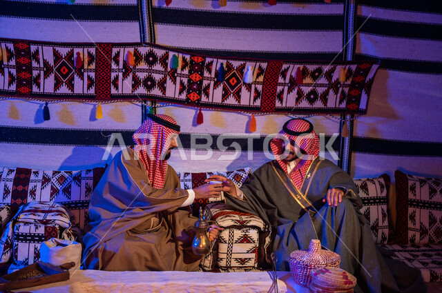 مفهوم حسن الضيافه والكرم، جلسات شعبية داخل خيمة تراثية، عادات وتقاليد سعودية اصيلة في تقديم القهوة العربية، ارتداء الازياء الوطنية، صب القهوة باستخدام الدلة النحاسية، صديقان عربيان خليجيان سعوديان يرتديان الشماغ والبشت يقضيان اوقات ممتعة في كشتة بر شبابية