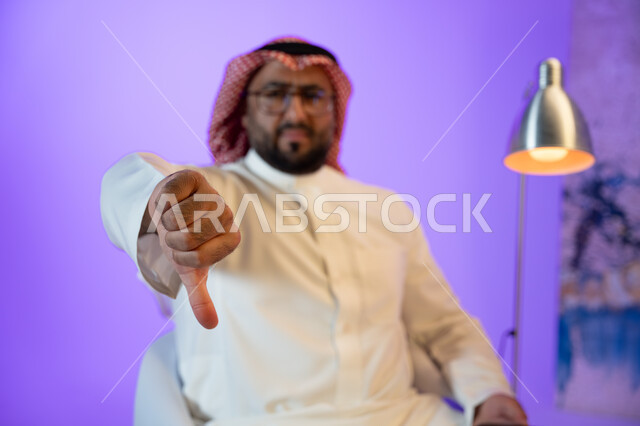Looking at the camera and pointing with his thumb down with a disliking gesture, a Saudi Gulf Arab man wearing a traditional thobe and shemagh, the concept of body language, movements and gestures