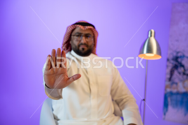 الجلوس في غرفة المعيشة والنظر إلى الكاميرا, اشارات يدين تدل على الأمر بالنهي عن فعل شيء ما, الصمت و التوقف عن الكلام، رجل عربي خليجي سعودي يرتدي الثوب والشماغ التقليدي يقوم برفع كفه للأعلى بإيماءات عدم الإعجاب