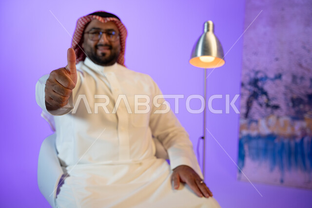 مفهوم لغة الجسد والحركات والاشارات, رجل عربي خليجي سعودي مبتسم يرتدي الثوب والشماغ التقليدي, النظر إلى الكاميرا والاشارة بإصبع الابهام بإيماءات الاعجاب والموافقة والرضى, الاهتمام بالمظهر الخارجي