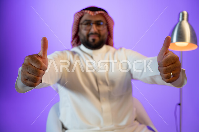 مفهوم لغة الجسد والحركات والاشارات, صورة مقربة لرجل عربي خليجي سعودي مبتسم يرتدي الثوب والشماغ التقليدي, النظر إلى الكاميرا والاشارة بإصبع الابهام بإيماءات الاعجاب والموافقة والرضى, الاهتمام بالمظهر الخارجي