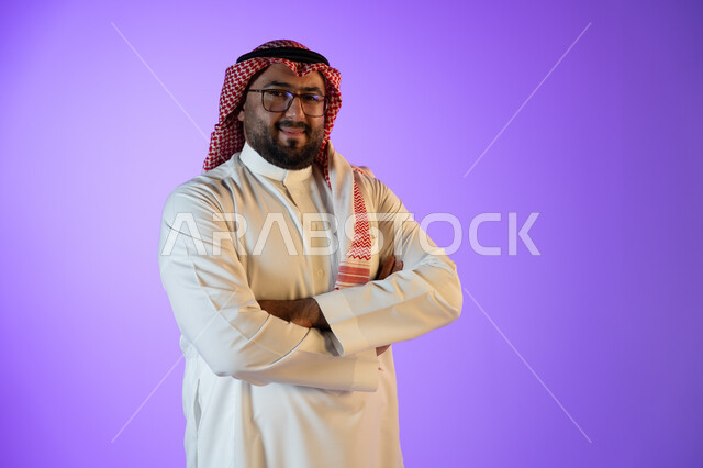 مفهوم الاهتمام بالمظهر الخارجي والشياكة, شخصيه سعوديه بارزه، النظر إلى الكاميرا بإيماءات أيدي متقاطعه والثقه والاعتزاز بالنفس، بورتريه لرجل عربي خليجي سعودي مبتسم يرتدي الثوب والشماغ التقليدي, خلفية ملونة