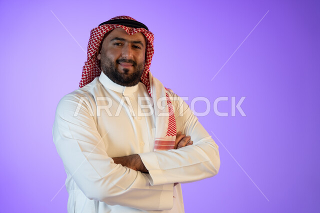 مفهوم الاهتمام بالمظهر الخارجي والشياكة, شخصيه سعوديه بارزه، النظر إلى الكاميرا بإيماءات أيدي متقاطعه والثقه والاعتزاز بالنفس، بورتريه لرجل عربي خليجي سعودي مبتسم يرتدي الثوب والشماغ التقليدي, خلفية ملونة