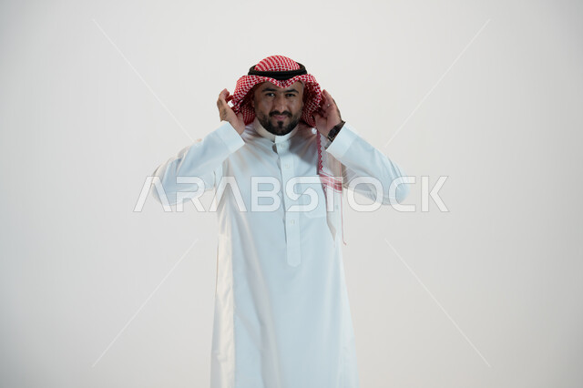 Looking at the camera, portrait of a Saudi Gulf Arab man wearing a traditional thobe and shemagh, placing his hands on his ear, listening to something with gestures of contemplation and concentration, the concept of body language and movements, white back