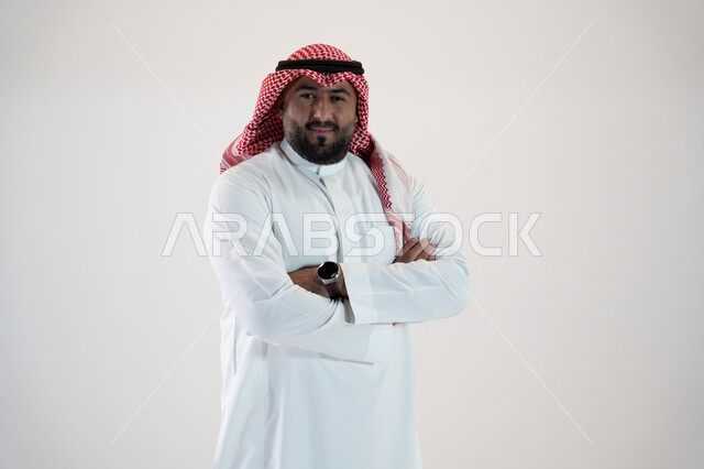 شخصيه سعوديه بارزه، النظر إلى الكاميرا بإيماءات أيدي متقاطعه والثقه والاعتزاز بالنفس، مفهوم الاهتمام بالمظهر الخارجي والشياكة ، بورتريه لرجل عربي خليجي سعودي مبتسم يرتدي الثوب والشماغ التقليدي, خلفية بيضاء