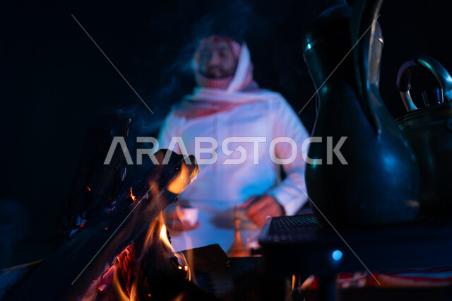 جلسة بر سعودية شعبية، شرب القهوة العربية, أجواء ليلية ممتعة, رجل عربي خليجي سعودي يرتدي الشماغ والزي التقليدي في رحلة تخييم برية في صحراء السعودية، صورة مقربة لدلة القهوة العربية مع شعلة حطب، كشتة بر شبابية مسائية