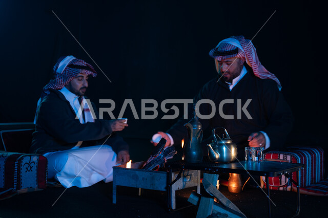 شرب القهوة العربية, أجواء شتوية ليلية ممتعة, رجلان عربيان خليجيان سعوديان يرتديان ملابس الشتاء الدافئة و يستمتعان بالتحدث في رحلة تخييم برية في صحراء السعودية، حوارات ونقاشات بين الأصدقاء، دلة القهوة العربية، جلسة بر سعودية شعبية، كشتة بر شبابية مسائية
