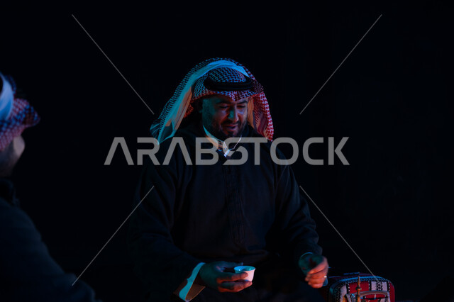 شرب القهوة العربية, أجواء شتوية ليلية ممتعة, رجلان عربيان خليجيان سعوديان يرتديان ملابس الشتاء الدافئة و يستمتعان بالتحدث في رحلة تخييم برية في صحراء السعودية، حوارات ونقاشات بين الأصدقاء، دلة القهوة العربية، جلسة بر سعودية شعبية، كشتة بر شبابية مسائية