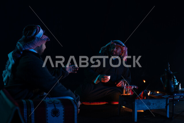 شرب القهوة العربية, أجواء شتوية ليلية ممتعة, رجلان عربيان خليجيان سعوديان يرتديان ملابس الشتاء الدافئة و يستمتعان بالتحدث في رحلة تخييم برية في صحراء السعودية، حوارات ونقاشات بين الأصدقاء، دلة القهوة العربية، جلسة بر سعودية شعبية، كشتة بر شبابية مسائية