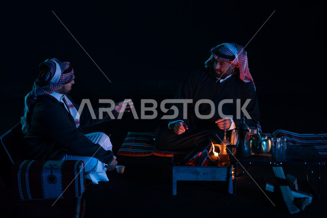 حوارات ونقاشات بين الأصدقاء، أجواء شتوية ليلية ممتعة, رجلان عربيان خليجيان سعوديان يرتديان ملابس الشتاء الدافئة و يستمتعان بالتحدث في رحلة تخييم برية في صحراء السعودية، دلة القهوة العربية، جلسة بر عربية شعبية، كشتة بر شبابية مسائية