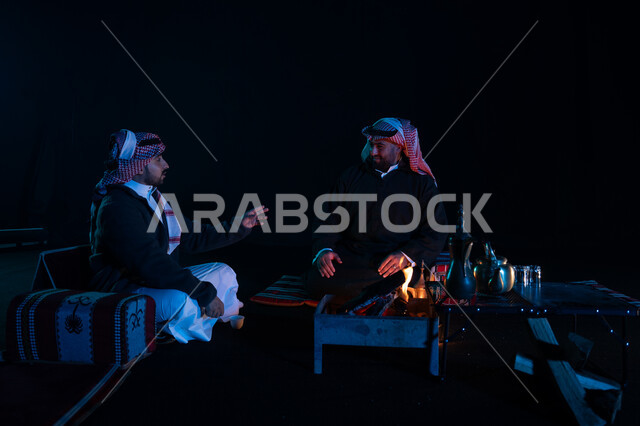 أجواء شتوية ليلية ممتعة, رجلان عربيان خليجيان سعوديان يرتديان ملابس الشتاء الدافئة و يستمتعان بالتحدث في رحلة تخييم برية في صحراء السعودية، حوارات ونقاشات بين الأصدقاء، دلة القهوة العربية، جلسة بر عربية شعبية، كشتة بر شبابية مسائية