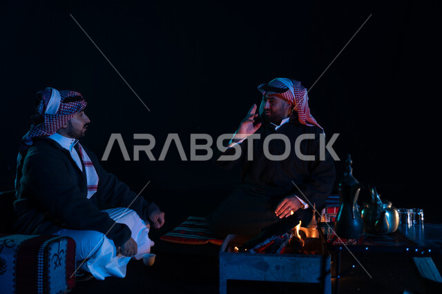أجواء شتوية ليلية ممتعة, رجلان عربيان خليجيان سعوديان يرتديان ملابس الشتاء الدافئة و يستمتعان بالتحدث في رحلة تخييم برية في صحراء السعودية، حوارات ونقاشات بين الأصدقاء، دلة القهوة العربية، جلسة بر عربية شعبية، كشتة بر شبابية مسائية