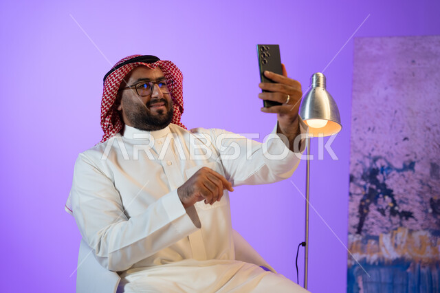 The concept of family ties, communicating with friends and relatives with gestures of pleasure, a Saudi Gulf Arab young man wearing a thobe and red shemagh making a video call via mobile phone, friendly chats and dialogues with friends, sitting and using