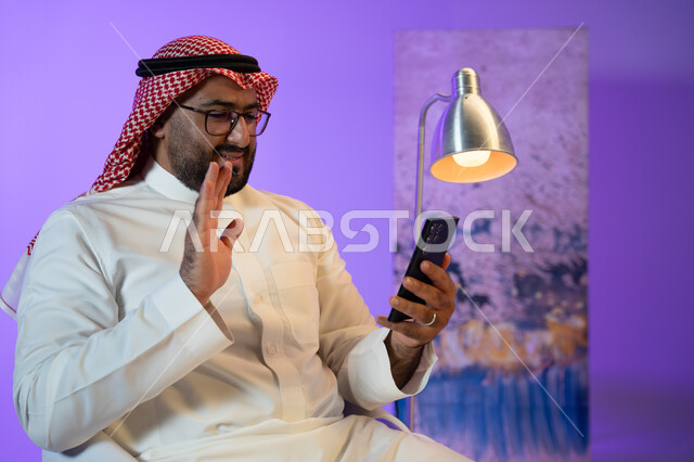 التواصل مع الأصدقاء والأقارب بإيماءات السرور, مفهوم صلة الرحم, شاب عربي خليجي سعودي يرتدي الثوب والشماغ الأحمر يقوم بإجراء مكالمة فيديو عبر الجوال, دردشات وحوارات ودية مع الأصحاب, الجلوس واستخدام تطبيقات التواصل الاجتماعي