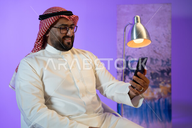 مفهوم صلة الرحم, التواصل مع الأصدقاء والأقارب بإيماءات السرور, شاب عربي خليجي سعودي يرتدي الثوب والشماغ الأحمر يقوم بإجراء مكالمة فيديو عبر الجوال, دردشات وحوارات ودية مع الأصحاب, الجلوس واستخدام تطبيقات التواصل الاجتماعي