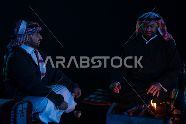 كشتة بر شبابية مسائية, حوارات ونقاشات بين الأصدقاء، أجواء شتوية ليلية ممتعة, رجلان عربيان خليجيان سعوديان يرتديان ملابس الشتاء الدافئة و يستمتعان بالتحدث في رحلة تخييم برية في صحراء السعودية، دلة القهوة العربية، جلسة بر عربية شعبية