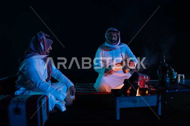 تقديم القهوة العربية للضيوف، الجلسات السعودية التقليدية الشعبية، قضاء أوقات ممتعة في الأجواء الليلية في الصحراء السعودية, رجلان عربيان خليجيان سعوديان يرتديان الشماغ والزي التقليدي في رحلة تخييم برية، كشتة بر شبابية مسائية, حسن الضيافة والاستقبال