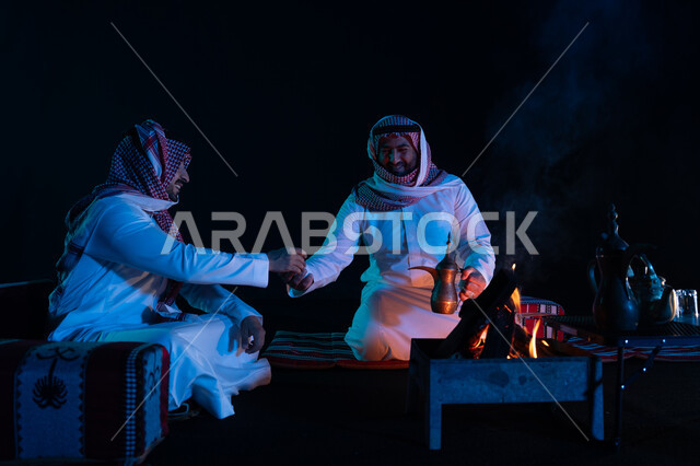 تقديم القهوة العربية للضيوف، الجلسات السعودية التقليدية الشعبية، قضاء أوقات ممتعة في الأجواء الليلية في الصحراء السعودية, رجلان عربيان خليجيان سعوديان يرتديان الشماغ والزي التقليدي في رحلة تخييم برية، كشتة بر شبابية مسائية, حسن الضيافة والاستقبال