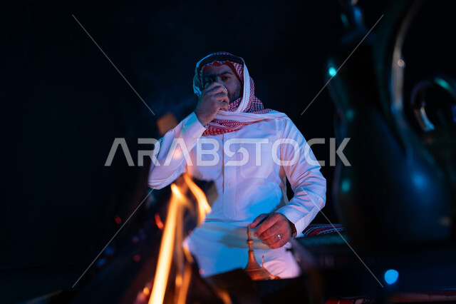 الاستمتاع بشرب القهوة العربية, صورة مقربة لدلة قهوة مع شعلة حطب في الصحراء السعودية، الجلسات السعودية التقليدية الشعبية، قضاء أوقات ممتعة في الأجواء الليلية, رجل عربي خليجي سعودي يرتدي الشماغ والزي التقليدي في رحلة تخييم برية، كشتة بر شبابية مسائية