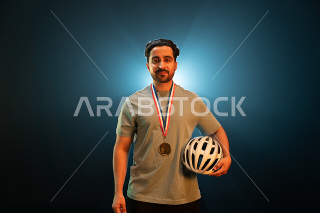 الفوز بسباق الدراجات الهوائية، إيماءات تدل على السعادة بتحقيق المركز الأول في المسابقة، بورتريه مقرب لشاب عربي خليجي سعودي ينظر الى الكاميرا يرتدي الميدالية الذهبية ويحمل الى جانبه خوذة الحماية، المحافظة على اللياقة الجسدية، خلفية سوداء مع إضاءة بيضاء