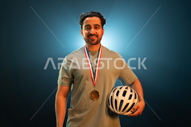 الفوز بسباق الدراجات الهوائية، إيماءات تدل على السعادة بتحقيق المركز الأول في المسابقة، بورتريه مقرب لشاب عربي خليجي سعودي ينظر الى الكاميرا يرتدي الميدالية الذهبية ويحمل الى جانبه خوذة الحماية، المحافظة على اللياقة الجسدية، خلفية سوداء مع إضاءة بيضاء