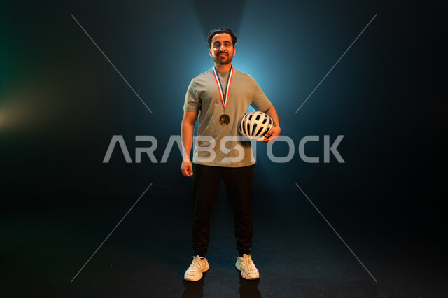 إيماءات تدل على السعادة بالحصول على المركز الأول في المسابقة، بورتريه لشاب عربي خليجي سعودي ينظر الى الكاميرا يرتدي الميدالية الذهبية ويحمل الى جانبه خوذة الحماية، الفوز بسباق الدراجات الهوائية، المحافظة على اللياقة الجسدية، خلفية سوداء مع إضاءة بيضاء