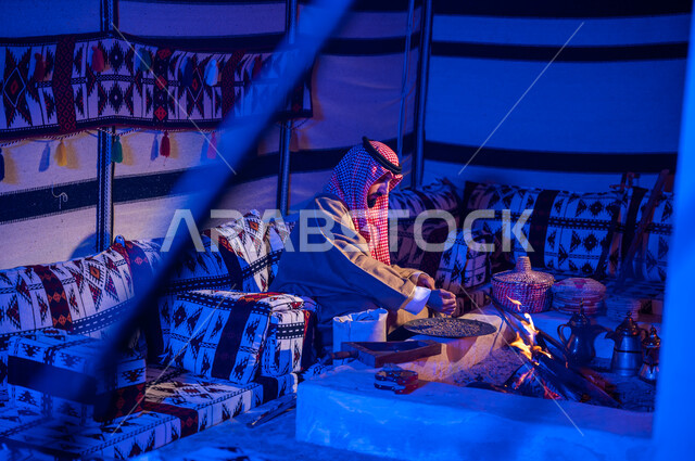 وضع حبوب البن في المحماس لتحميصها على شعلة النار، رجل عربي خليجي سعودي يرتدي الشماغ والبشت يستخدم المعاميل لإعداد القهوة بالطريقة التقليدية على الحطب في موقد طيني داخل خيمة شعبيه تراثية، منتج محلي ومحاصيل زراعية وطنية، أسلوب الحياة القديمه في الباديه