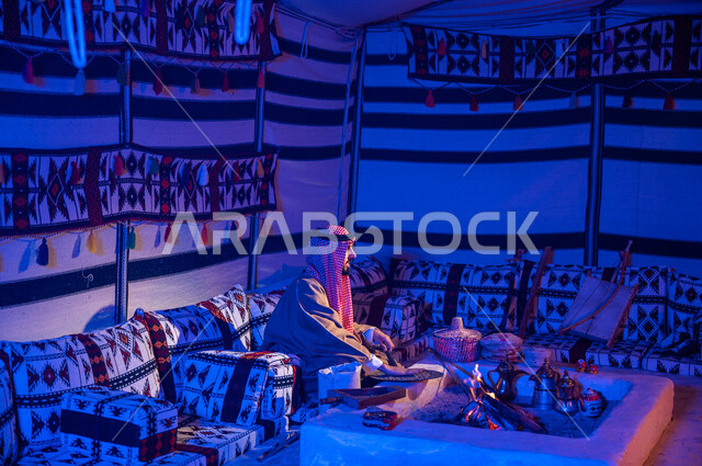 وضع حبوب البن في المحماس لتحميصها على شعلة النار، رجل عربي خليجي سعودي يرتدي الشماغ والبشت يستخدم المعاميل لإعداد القهوة بالطريقة التقليدية على الحطب في موقد طيني داخل خيمة شعبيه تراثية، منتج محلي ومحاصيل زراعية وطنية، أسلوب الحياة القديمه في الباديه