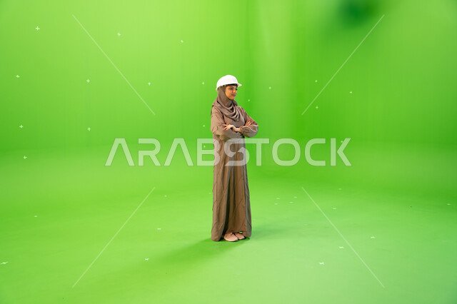 Standing with crossed hands, female engineers in the Kingdom of Saudi Arabia, managing and studying engineering projects, portrait of a veiled Saudi Arabian Gulf engineer wearing an abaya and a protective helmet with gestures indicating confidence, full-l