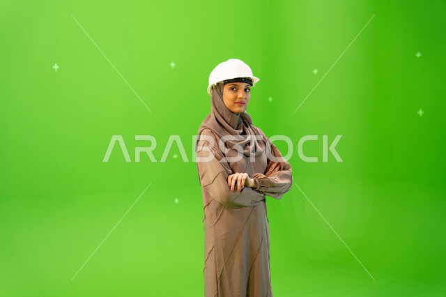 مهندسات المملكة العربية السعودية, إدارة ودراسة المشاريع الهندسية، بورتريه من الجانب لمهندسة عربية خليجية سعودية محجبة ترتدي العباءة و خوذة الحماية تنظر الى الكاميرا بإيمات تدل على الثقة، الوقوف بأيدي متقاطعة، كروما، خلفية خضراء