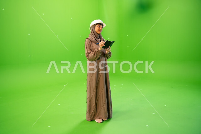 تقييم البيانات الإنشائية في موقع العمل, بورتريه لمهندسة عربية خليجية سعودية ترتدي العباءة السعودية و خوذة الحماية وتقوم بكتابة وتدوين الملاحظات، إدارة ودراسة المشاريع الهندسية، الإشراف على العمل وتدقيقه، صورة الجسم بالطول الكامل, خلفية خضراء, كروما