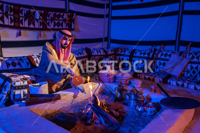Preparing the stove for heating in the winter weather. A Saudi Gulf Arab man wearing a shemagh and bisht uses the popular pump to light the fire and roast the coffee beans. Enjoy the popular gatherings inside a heritage tent. Follow Saudi customs and traditions in preparing Arabic coffee.