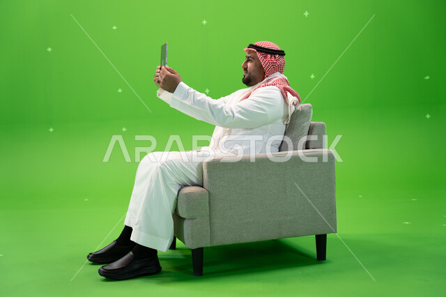 Sharing happy moments on social networking sites, taking souvenir selfies using a tablet, using modern technical devices, a portrait from the side of a Saudi Gulf Arab man wearing a shemagh and traditional dress, sitting in a comfortable chair and using a