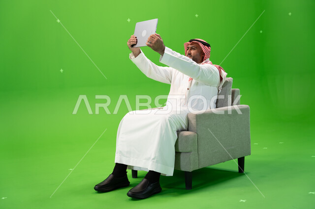 مشاركة اللحظات السعيدة على مواقع التواصل الإجتماعي, التقاط صور سيلفي تذكارية باستخدام التابلت, استعمال أجهزة تقنية حديثة، بورتريه من الجانب لرجل عربي سعودي خليجي يرتدي الشماغ و الزي التقليدي يجلس على كرسي مريح ويستخدم الجهاز اللوحي، خلفية خضراء, كروما
