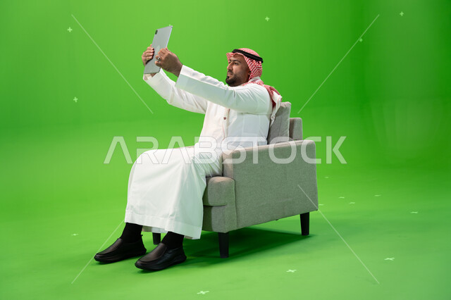 Sharing happy moments on social networking sites, taking souvenir selfies using a tablet, using modern technical devices, a portrait from the side of a Saudi Gulf Arab man wearing a shemagh and traditional dress, sitting in a comfortable chair and using a