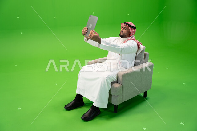 التقاط صور سيلفي تذكارية باستخدام التابلت, مشاركة اللحظات السعيدة على مواقع التواصل الإجتماعي, استعمال أجهزة تقنية حديثة، بورتريه من الجانب لرجل عربي سعودي خليجي يرتدي الشماغ و الزي التقليدي يجلس على كرسي مريح ويستخدم الجهاز اللوحي، خلفية خضراء, كروما