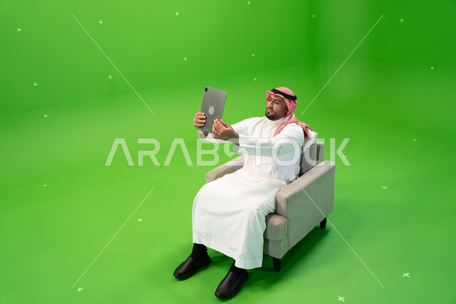التقاط صور سيلفي تذكارية باستخدام التابلت, مشاركة اللحظات السعيدة على مواقع التواصل الإجتماعي, استعمال أجهزة تقنية حديثة، بورتريه من الأعلى لرجل عربي سعودي خليجي يرتدي الشماغ و الزي التقليدي يجلس على كرسي مريح ويستخدم الجهاز اللوحي، خلفية خضراء, كروما