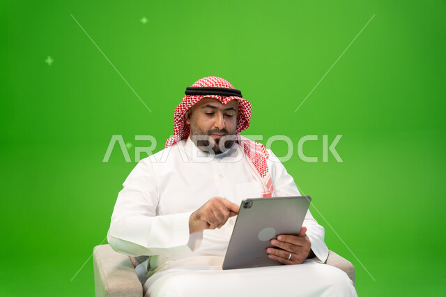 التسوق عبر الانترنت باستخدام التابلت, قضاء الوقت بتصفح برامج التواصل الاجتماعي, إنجاز المهام عن بعد، استعمال أجهزة تقنية حديثة، بورتريه لرجل عربي سعودي خليجي يرتدي الشماغ و الزي التقليدي يجلس على كرسي مريح ويستخدم الجهاز اللوحي، خلفية خضراء, كروما