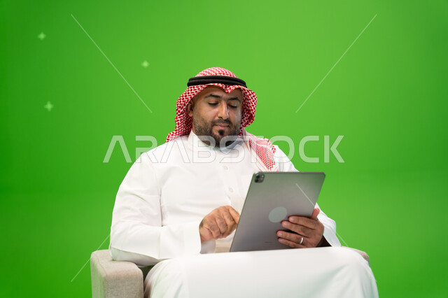 Completing tasks remotely, spending time browsing social media programs, shopping online using a tablet, using modern technical devices, a close-up portrait of a Saudi Gulf Arab man wearing a shemagh and traditional dress, sitting in a comfortable chair a