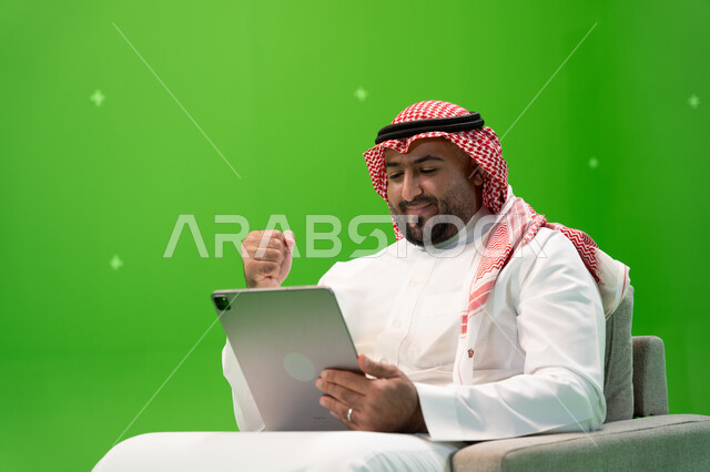 إيماءات  تدل على السعادة بالفوز وتحقيق الأهداف, القيام بمكالمة عمل ، التحدث مع الأهل و الأصدقاء عن بعد, بورتريه مقرب من الجانب لرجل عربي سعودي خليجي يرتدي الزي التقليدي يجلس على الكرسي ويقوم بإجراء مكالمة فيديو عبر التابلت، خلفية خضراء, كروما