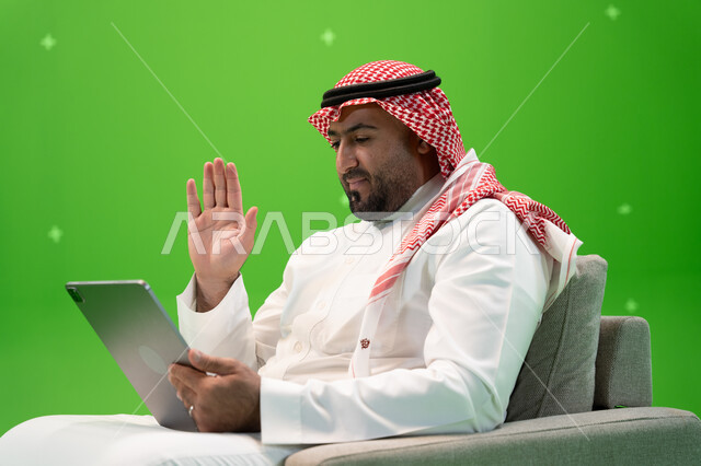 التحدث مع الأهل و الأصدقاء عن بعد, إيماءات يدين تدل على الترحيب, محادثات ودردشات عبر الانترنت, القيام بمكالمة عمل ، بورتريه مقرب من الجانب لرجل عربي سعودي خليجي يرتدي الزي التقليدي يجلس على الكرسي ويقوم بإجراء مكالمة فيديو عبر التابلت، خلفية خضراء, كروما