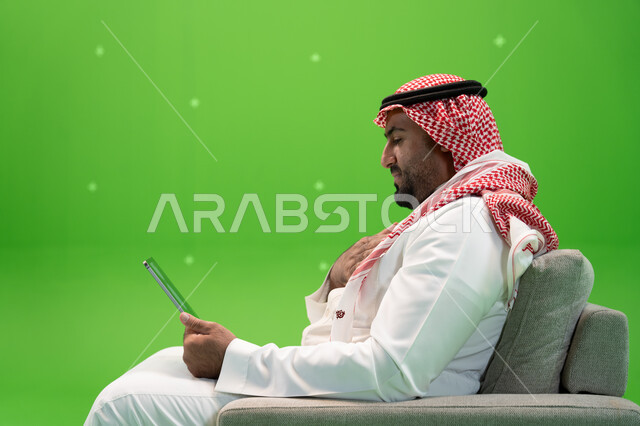 القيام بمكالمة عمل ، محادثات ودردشات عبر الانترنت, التحدث مع الأهل و الأصدقاء عن بعد, بورتريه مقرب من الجانب لرجل عربي سعودي خليجي يرتدي الزي التقليدي يجلس على الكرسي ويقوم بإجراء مكالمة فيديو عبر التابلت، خلفية خضراء, كروما