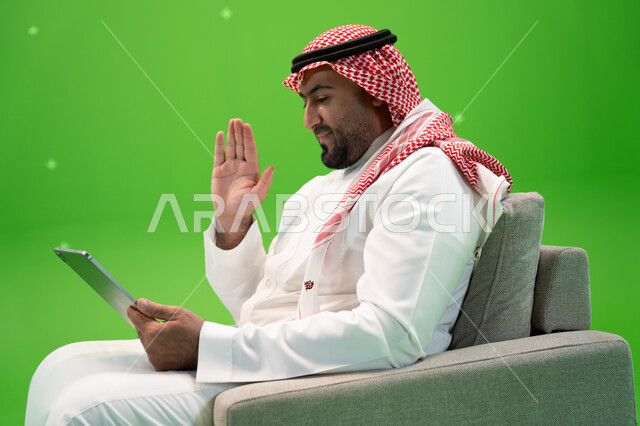 محادثات ودردشات عبر الانترنت, إيماءات يدين تدل على الترحيب, القيام بمكالمة عمل ، التحدث مع الأهل و الأصدقاء عن بعد, بورتريه مقرب من الجانب لرجل عربي سعودي خليجي يرتدي الزي التقليدي يجلس على الكرسي ويقوم بإجراء مكالمة فيديو عبر التابلت، خلفية خضراء, كروما