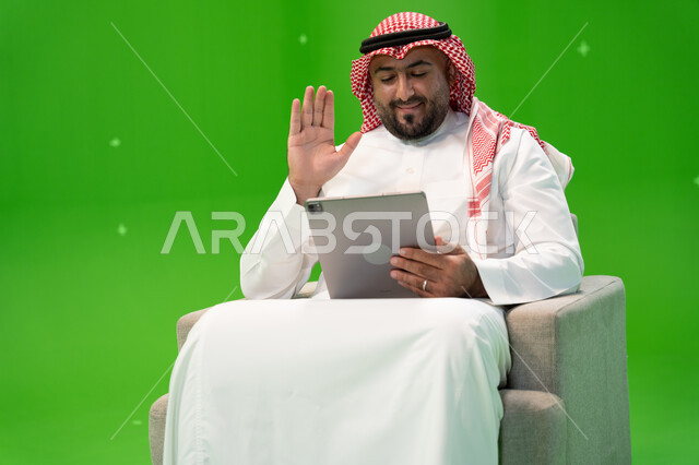 إيماءات يدين تدل على الترحيب, محادثات ودردشات عبر الانترنت, القيام بمكالمة عمل ، التحدث مع الأهل و الأصدقاء عن بعد, بورتريه  لرجل عربي سعودي خليجي يرتدي الزي التقليدي يجلس على الكرسي ويقوم بإجراء مكالمة فيديو عبر التابلت، خلفية خضراء, كروما