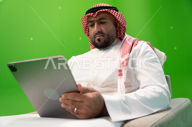Spending time browsing social media programs, completing tasks remotely, shopping online using a tablet, using modern technical devices, a close-up portrait of a Saudi Gulf Arab man wearing a shemagh and traditional dress, sitting in a comfortable chair a