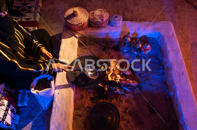 وضع حبوب البن في المحماس لتحميصها على شعلة النار في موقد طيني، إمرأه عربيه خليجية سعودية ترتدي العباءة والبرقع تستخدم المعاميل لإعداد القهوة بالطريقة التقليدية على الحطب داخل خيمة شعبيه تراثية، منتج محلي ومحاصيل زراعية وطنية، أسلوب الحياة القديمه في الباديه
