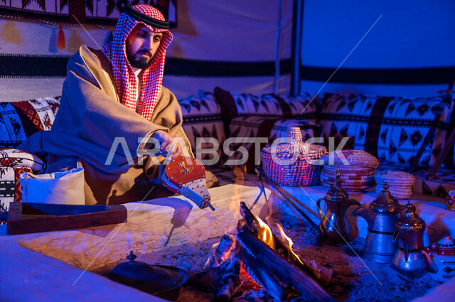 تجهيز الموقد للتدفئة في الاجواء الشتوية, رجل عربي خليجي سعودي يرتدي الشماغ والبشت يقوم باستعمال المضخة الشعبية لإشعال النار وتحميص حبوب البن, الاستمتاع بالجلسات الشعبية داخل خيمة تراثية, اتباع العادات والتقاليد السعودية في تحضير القهوة العربية